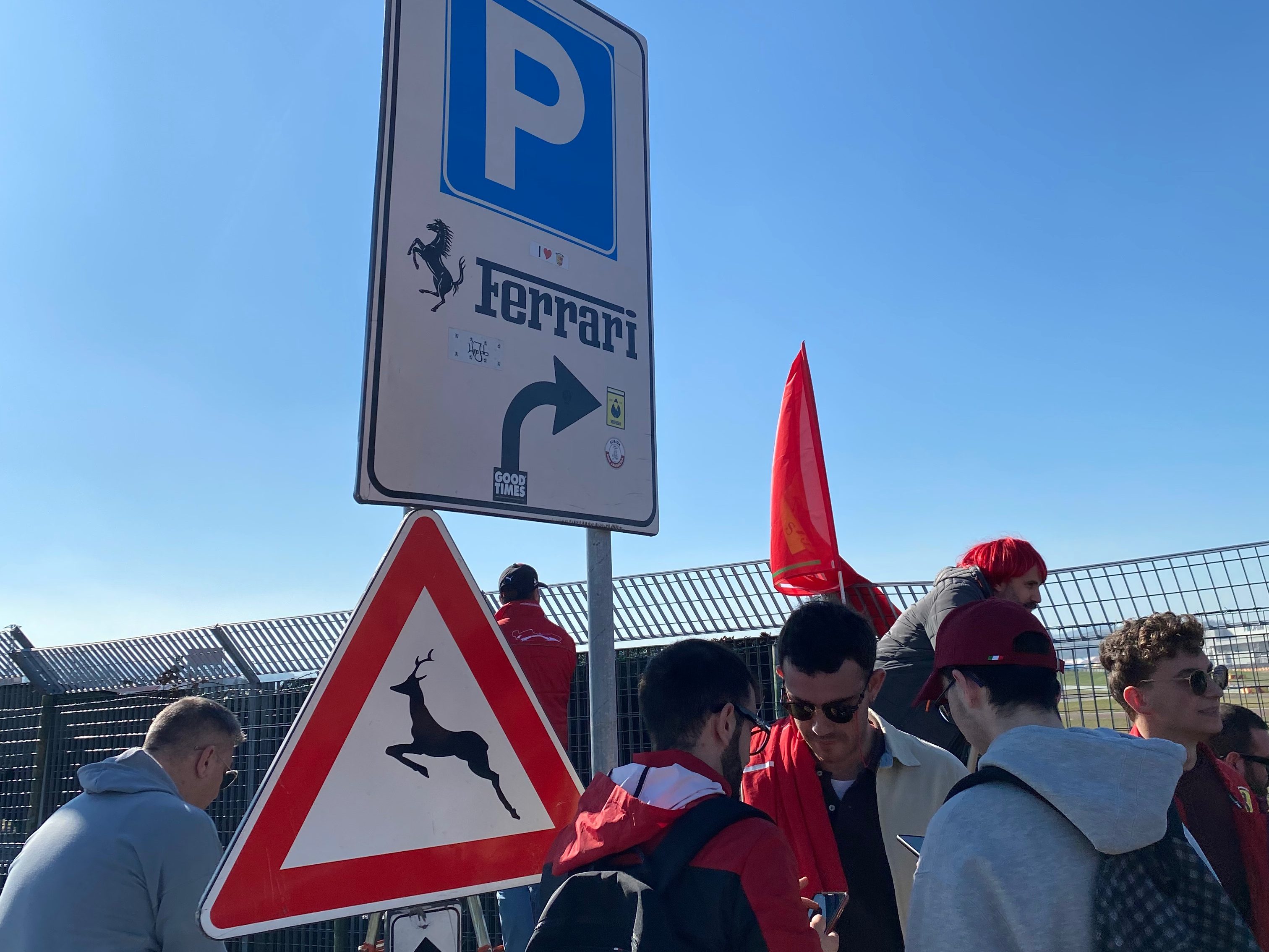 Cartello di parcheggio Ferrari con un simbolo di avvertimento per il passaggio di animali, con tifosi Ferrari in abbigliamento rosso sullo sfondo.